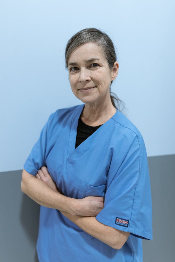 Middle-aged nurse smiling confidently in blue scrubs, crossed arms in a clinic setting.